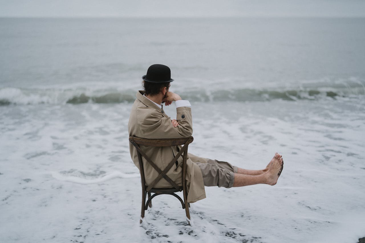 Man in coat and hat sitting on a chair at the edge of the ocean, lost in thought.